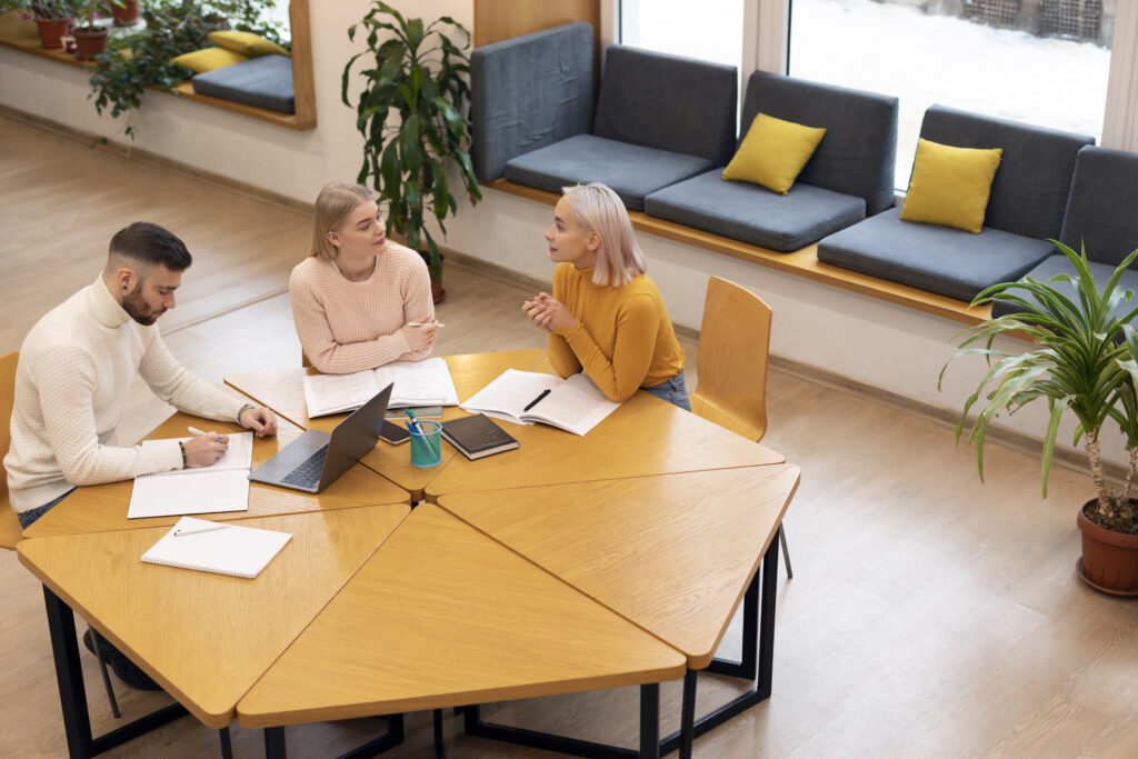 Three friends are reading books and notes at a library table, illustrating different office desk materials like laminate, wood, and glass.