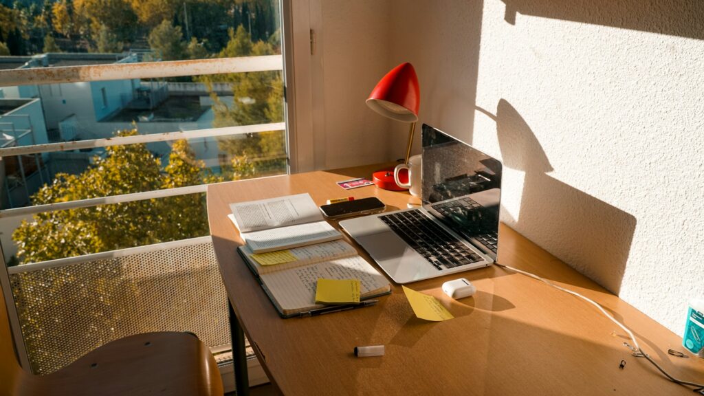Modern home office desk with a laptop, notebook, and office supplies near a bright window.