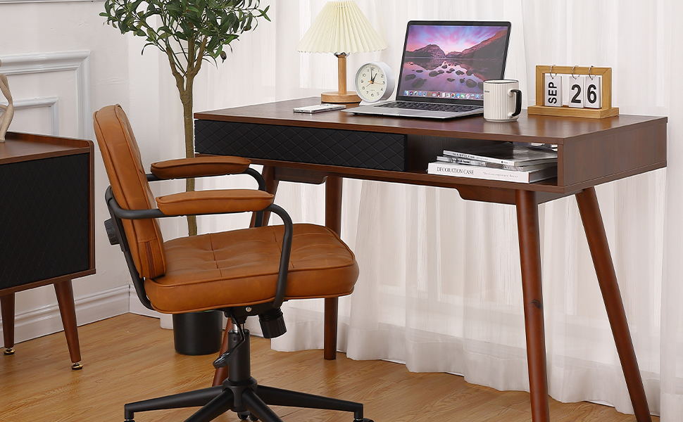 Mid-century modern wooden desk with a laptop, table lamp, clock, and calendar, paired with a brown leather office chair in front of sheer curtains.